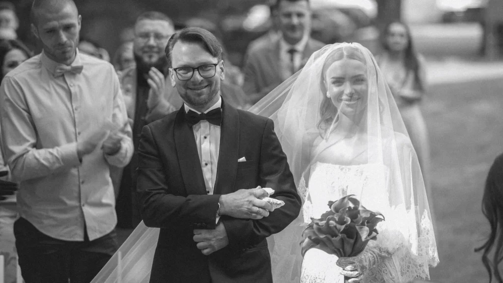 Panna młoda prowadzona do ołtarza w ogrodzie, goście uśmiechają się i klaszczą