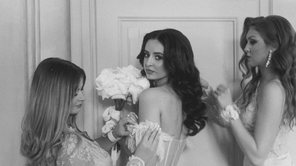 Bridesmaids lacing the bride into her gown, white rose bouquet in hand