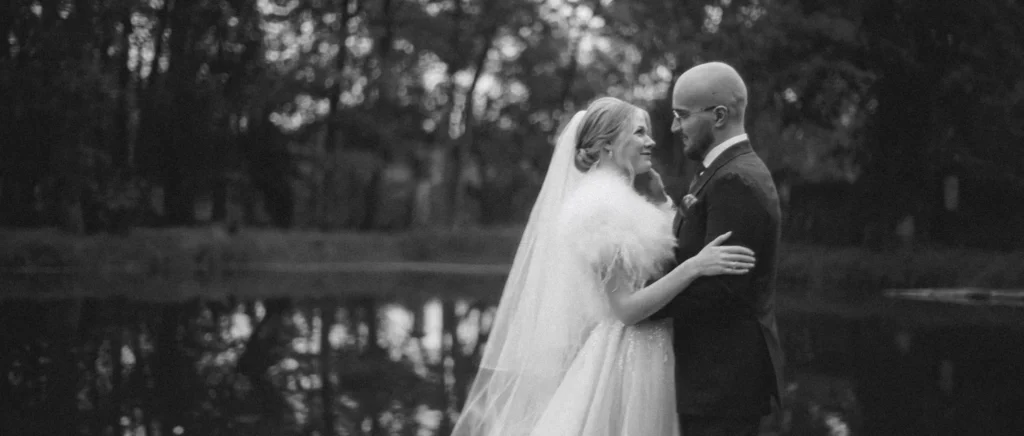 Newlyweds embracing by a pond, the bride's veil trailing behind her